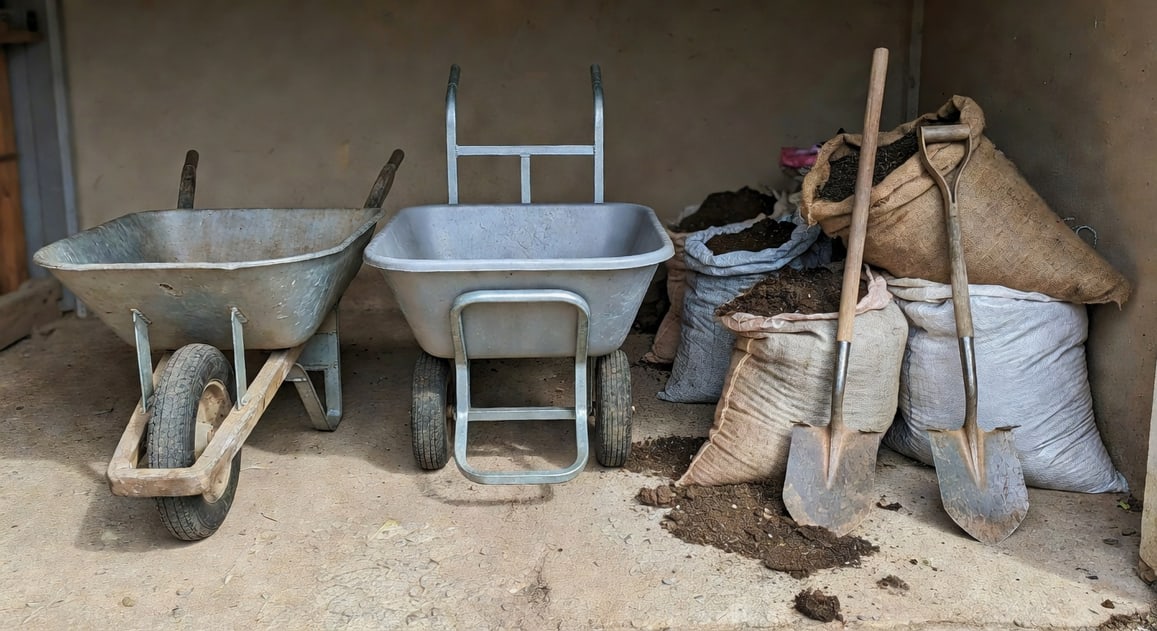 工具間並排鋼槽木把手獨輪車和塑膠槽鋼把手雙輪手推車，旁邊堆土袋與鏟子。